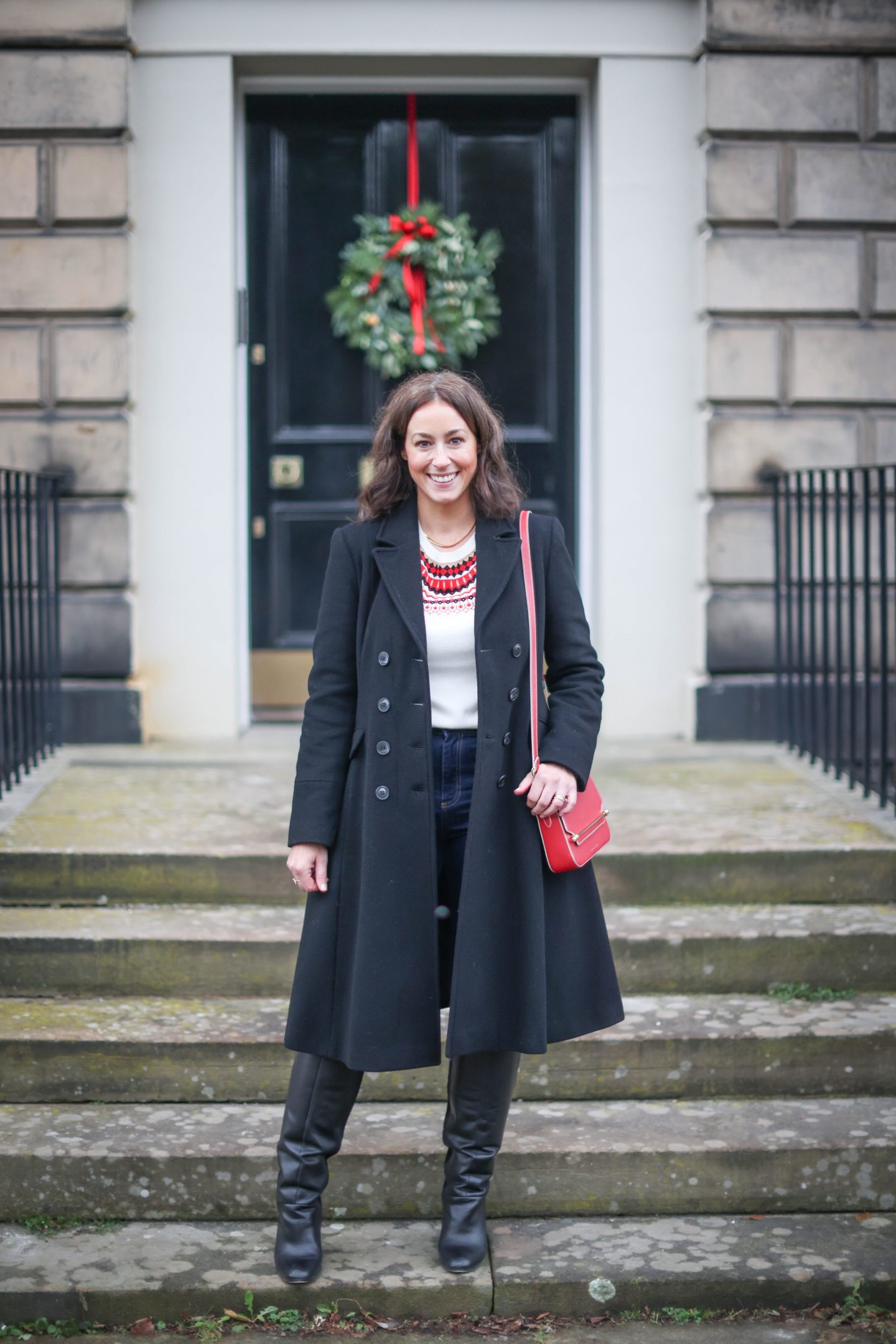 A woman wearing a fit and flare black coat in front of a door with a Christmas wreath