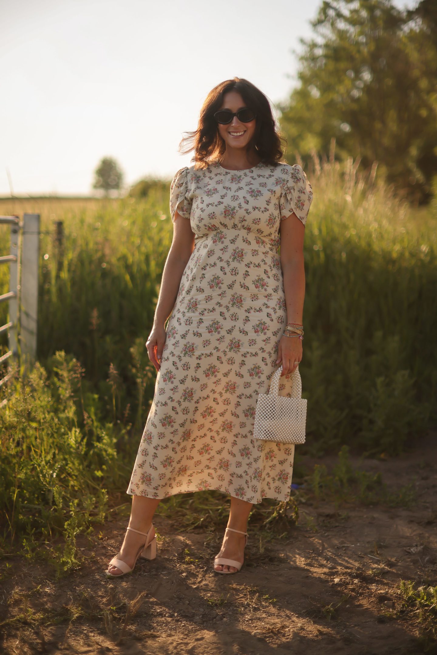 Christina stands beside a field of long grass wearing a summer dress and sunglasses as the sun sets.