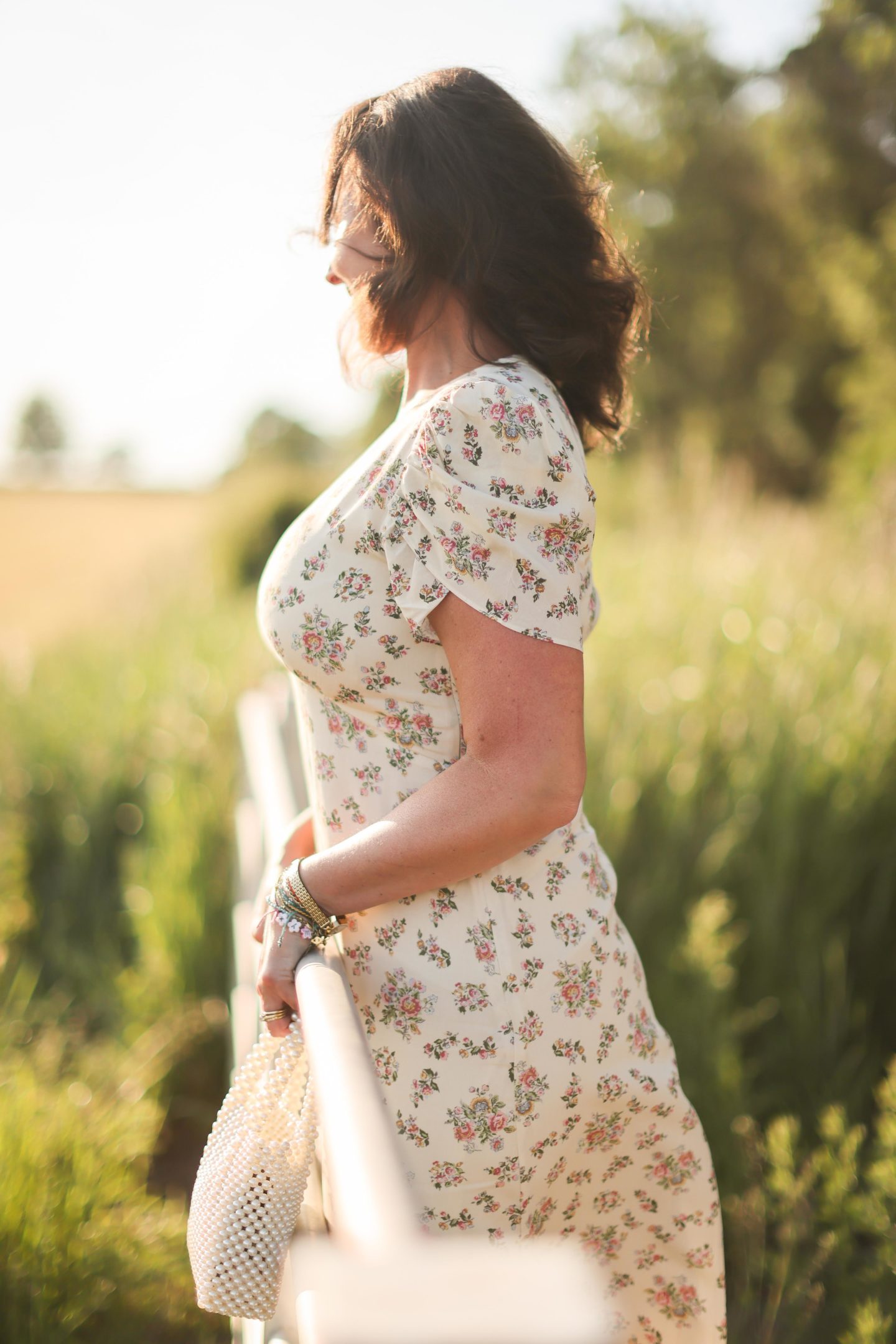 Christina stands beside a field of long grass wearing a summer dress looking away from camera as the sun sets.